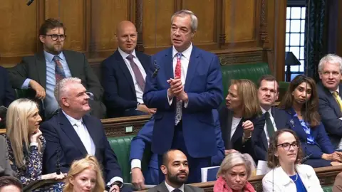 House of Commons/UK Parliament/PA Wire Nigel Farage, wearing a dark blue suit, white shirt and red and blue patterned tie, stands in the House of Commons while surrounded by other MPs