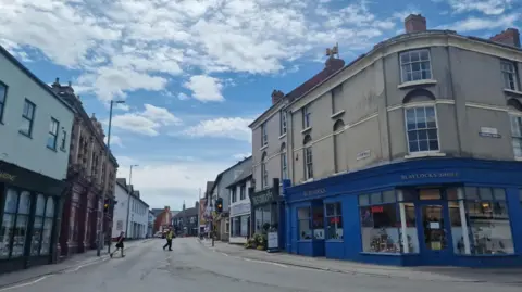 A road through Old Town, in Swindon. Two people are crossing the road. Commercial buildings line the streets on both sides of the carriageway.