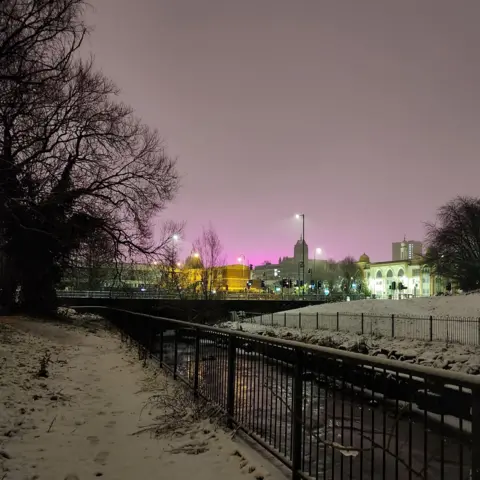 EliP/BBC Weather Watchers A river is in the foreground and lit up buildings can be seen behind. The sky is strange tint of pink and purple.