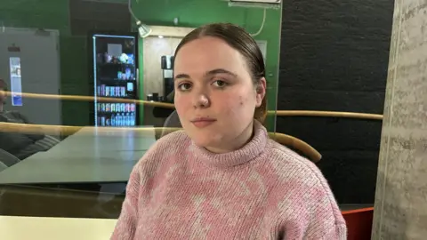 Amadea Topalli, a young woman with her hair pulled back, wearing a pink and white knitted jumper, looking into the camera.