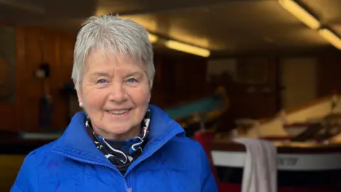 An elderly lady is stood with short grey hair and blue jacket on she has pearl earrings and a navy scarf on. She is stood in front of two St Ayles skiffs, one has a towel draped over the tip of it.