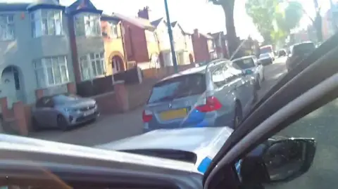 A blue car in an urban street seen from the door of a police vehicle