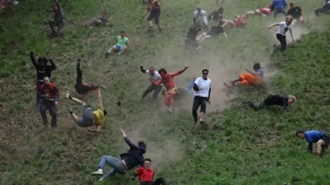 Image shows men running and tumbling down Cooper's Hill. The hill is very steep and grassy. The ground is so dry that there are dust trails behind the men as they run. Mot of the men have fallen over and are rolling and falling down the hill, with only a few of them still on their feet.