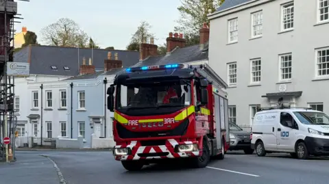 Guernsey Fire and Rescue Service A fire engine is parked across a road that is lined with buildings. There is a car and a van parked along the road. There is scaffolding on a building to the left.