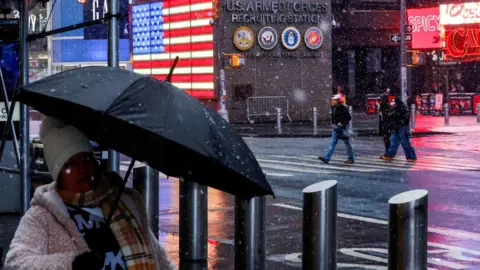 People walk through Times Square, wearing scarves hats and one is holding an umbrells as snow falls during a winter storm in New York City on 22 February 2026.
