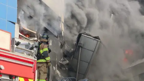 A firefighter stands in front of a building as smoke and flames billow