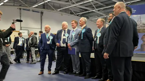 Eight men stand in a line in front of the stage and smile for the photographer in front of them. They are all wearing turquoise rosettes for Reform.