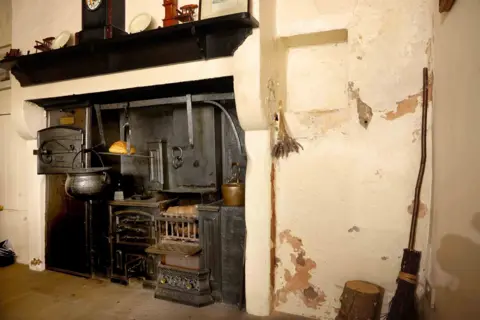 Colin Hattersley An old-fashioned fireplace with a pot hanging down from it, white walls and a wooden brush in the corner