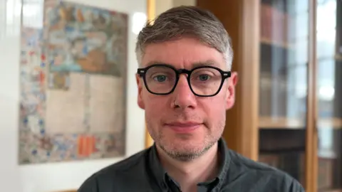 Chris Cassels, a man with short greying hair and glasses, standing in front of a bookcase and a wall chart.