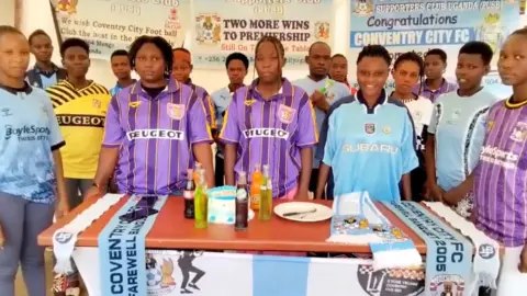 A group of men and women in football T-shirts stood around a table with Sky Blues merchandise, a cake and drinks