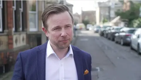 Alex Beckett with short dark hair and beard looking to the right of the camera wearing a blue jacket and light-coloured shirt. He is standing with a residential street behind him with parked cars to his left.