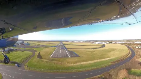 A view from beneath the wing of a small plane as it comes into land at Gloucestershire Airport. It is flying over a quiet road and heading straight for the runway. There are a few roads on the airfield itself, which is surrounded by countryside. A few warehouses, hangars and houses can be seen in the distance.