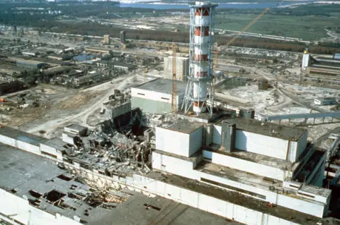 Getty Images A nuclear power plant, including a large, white, utilitarian building topped by a tall red and white circular chimney. Severe damage is visible to part of the structure, which appears to have been blown away, leaving twisted metal and a missing roof. Green fields, trees and a lake can be seen in the background. 