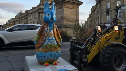 St Oswald's Hospice A Peter Rabbit statue which has been painted blue with an orange coat and painted body. The body is painted into separate lines with waves, flowers and the sea filling each one. In is standing in front of the base of Grey's Monument and a white car is driving behind it. There is a yellow and black forklift to the right of the sculpture.