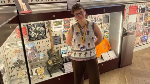 Beth stands in front of a guitar, and merchandise includes Radiohead South Park tickets.