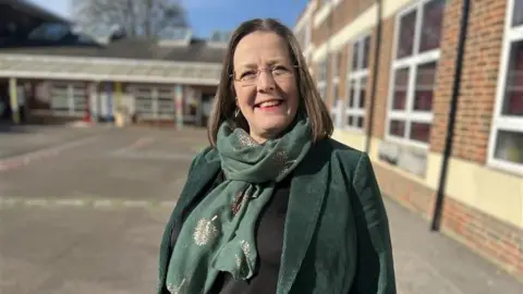 Headteacher Jo Reid is standing in a school playground wearing a green jacket and scarf. She has shoulder-length brown hair.