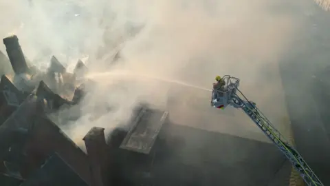 Stephen Barnes  (Bowline Images) Drone footage from above of a red-brick building with flames inside it. The building is large and long with its roof caved in. A firefighters in hi-viz protective clothing is on an aerial platform spraying it with water. 