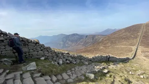 NI Water The Mourne wall seen on a sunny day. It's a wide shot that takes in a long, undulating portion of the wall, and some damage is visible