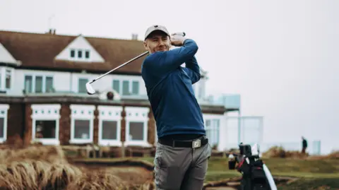 Golf England Golfer Josh Simpson stood with his club after teeing off with a clubhouse behind him in the distance.