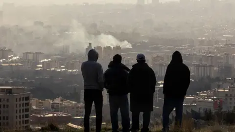 Getty Images Men watch from a hillside as a plume of smoke rises after an explosion in Tehran, Iran. Beneath them lies the city.