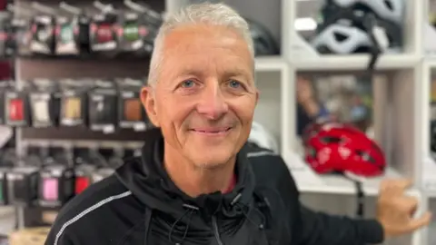 A portrait image of Paul Stewart, mechanic at The Bike Lounge in Beeston. He is stood in the shop in front of a wall of helmets.