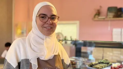 A woman stands in front of a bakery counter with an assortment of cakes and pastries. She wears a white hijab, brown glasses, a grey and white striped jumper and a brown apron with the word "Helmi's" on it. 