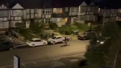 A biker doing a wheelie at night-time in a residential terraced street with cars parked along the roadsides.