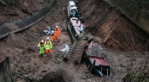 A muddy trench with three narrow boats in the mud and four people in reflective jackets