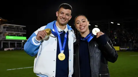 Patrick Khachfe/Onside Images Matt Weston and Tabby Stoecker hold up their gold medals at the Recreation Ground in Bath. He is wearing a dark shirt underneath a white jersey jacket with light blue sleeves and collar. She is wearing a black top underneath a black leather jacket. They are standing on a green pitch in the dark, but the light is bright on their faces.