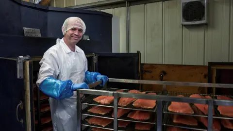 A man wearing a hairnet and white overalls holding a trolley on which there are several trays of salmon.