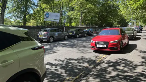 Aisha Iqbal/BBC A road outside a school with the school sign in the background. Cars lining the street on both sides, many of them parked on double yellow lines and across the pavement.
