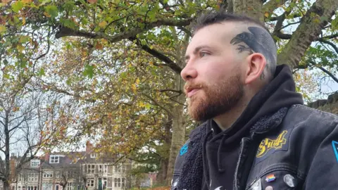 A side-profile image of Patrick Darling, from Bristol. He has short spikey hair, which is shaved at the sides. He has a tattoo on the left side of his head. He has a beard and a piercing in his left ear.