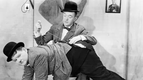Getty Images Stan Laurel sews the trousers of Oliver Hardy, who is bent over his knee, as they perform a scene in the film Hollywood Party.