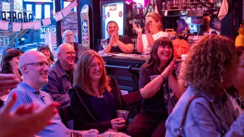 Jimmy Hill Photography A group of people are inside a pub and they are smiling and laughing. There are two women behind a dark brown wooden bar and they are clapping. The lighting is slightly dark and las pinkish red lights