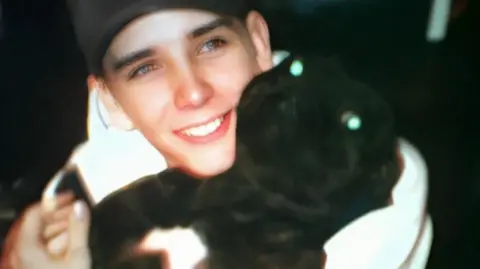 A young looking man is holding what appears to be a puppy in his arms. The man isn't looking directly at the camera, but he is smiling.