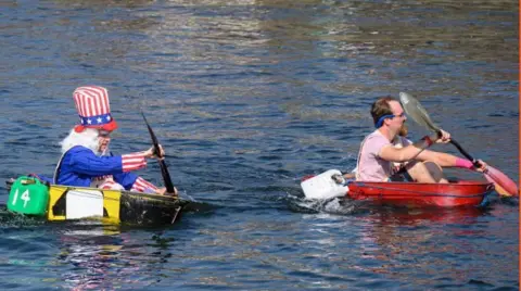 Two men paddling tin baths in the harbour, one is is fancy dress as Uncle Sam.