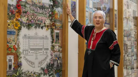 Leeds City Council Artist Kate Peace has short grey hair and is wearing a black and red dress. She is standing beside tapestry panels at Leeds Central Library