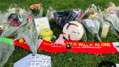 Tributes left in a park. Bunches of flowers have been left on the grass along with framed messages, a football, a scarf and cards.