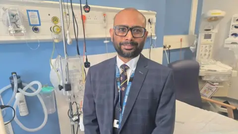 Landscape photo of Noel Peter, Director for Research, Innovation and Genomics at Gloucestershire Hospitals. He is standing by an empty hospital bed, surrounded by medical equipment. He is wearing a chequered suit and striped tie. He has black-framed, square glasses. 