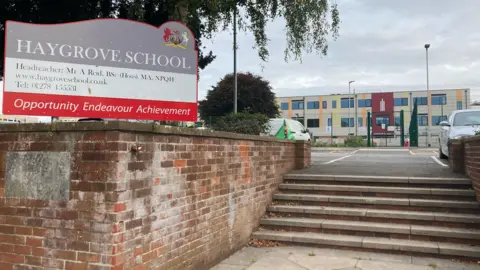 BBC A sign outside Haygrove School bearing the name of the school on a grey background with a motto in red reading: "Opportunity, endeavour, achievement". There is a car park behind and the school building in the distance, a modern three-tier unit with patches of red, yellow and orange cladding.