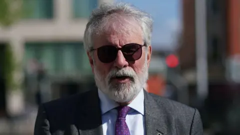 PA Media Gerry Adams, wearing a grey suit jacket, a light coloured collared shirt and purple tie. He has grey hair and a grey beard and wears dark sunglasses. He is walking on the street outside of the High Court, but the background behind him is blurred.