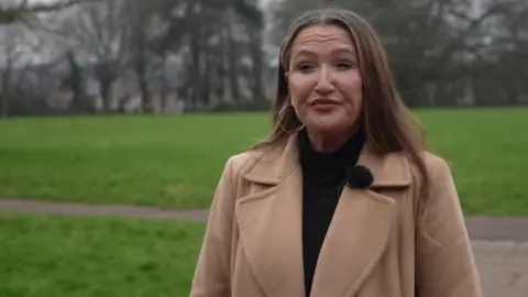 Kate Northcott Spall - a woman wearing a brown collared long coat standing outside in gardens.