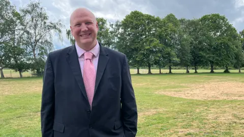 Clare Worden/BBC Headteacher at Reepham High Jon Croucher is wearing a black suit, pink shirt and pink tie. He is a bald man. He is smiling at the camera. He is standing in a grass field which is lined by tall trees in full leaf. This is where the new specialist resource base will be built. 