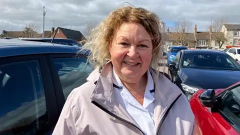 Vikki Irwin/BBC Hayley Foulger is wearing a lilac coat and white shirt underneath. She is standing in a car park with cars all around her. 