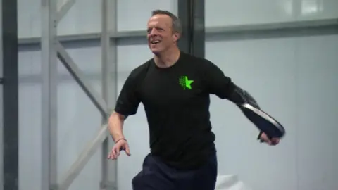 BBC Andy Knee playing padel, wearing a black t-shirt and tracksuit bottoms, holding a racquet.