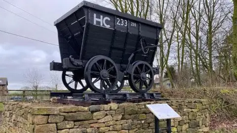 The Hetton Chaldron Wagon is a small black wooden carriage with four wheel and HC 233 written across it. It is placed on top of a stone fencing. There is a plaque beneath it. 