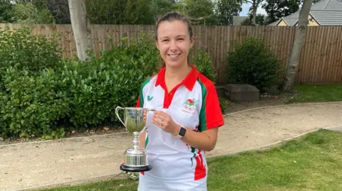 Chelsea Spencer. She is dressed in a white, red and green sports outfit and is holding a sports trophy in a garden. 