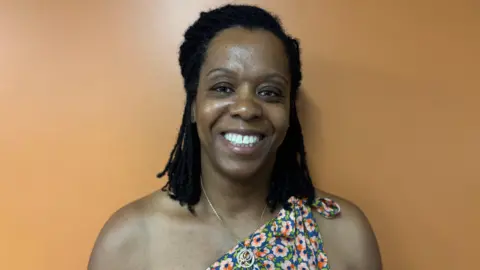 Amanda Toussaint smiling and wearing a flowery top in front of an orange wall.