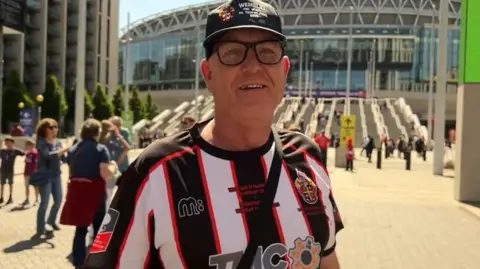Ian is outside Wembley wearing a cap and his football strip. He is smiling and there are lots of people behind him.