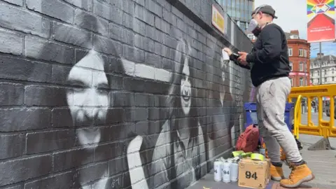 Mr Murals working on his mural in Birmingham. He is wearing a mask and side on to the camera, to the right on the photo.
